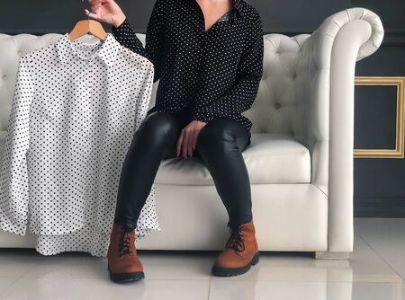 A girl in black leather trousers and suede boots demonstrates a white blouse with black polka dots. Without a face, sitting on a white leather sofaの写真素材