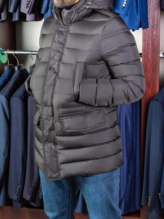 A young man is standing in a long jacket at a clothing store with his hands in his pockets. Assortment of goods in a boutique. Vertical layoutの写真素材