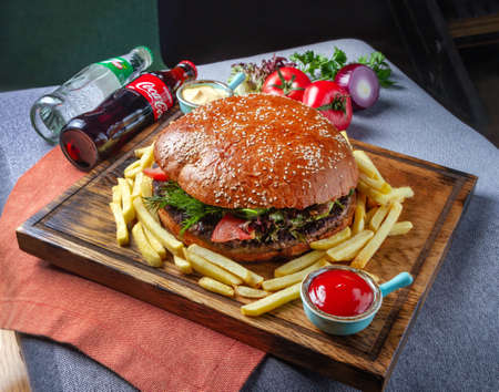 Huge burger on a serving board. Bottled carbonated drinks, fresh vegetables and herbs in the backgroundのeditorial素材