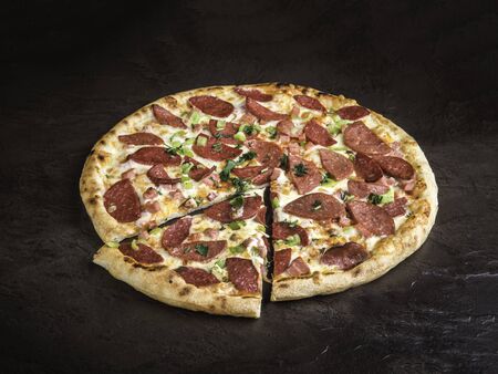 Pizza with two varieties of sausages, cheese and bell pepper on a dark background. A little sprinkled with fresh herbs. One piece is cut off and lies nearby. Close up, selective focus, side viewの写真素材