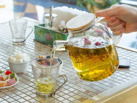 A hand holds a glass teapot by the handle, pouring herbal tea into a cup. Natural daylight, foreground focus, side viewの写真素材