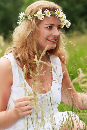 Young blond plus-size model in the white linen dress and flowers wreath sits laughing in a meadow in the high grassの写真素材