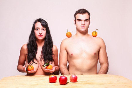 Conceptual image of a young nude man and woman posing with fresh tropical fruit in their hands and balanced on their shoulders looking at the camera with serious expressions as they sit at a tableの写真素材