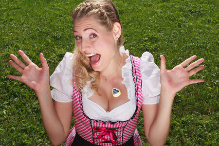 Fun loving carefree attractive young blond girl in a dirndl with a heart-shaped candy on her chest throwing up her hands and laughing, high angle view over green grass.の写真素材