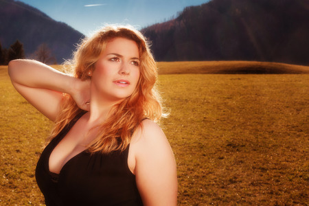 Portrait of a beautiful blonde woman with luscious cleavage in black tank dress. Illuminated on a meadow, the Alps in the background of the autumn sun.の写真素材