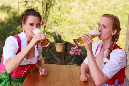 Two attractive Bavarian women wearing traditional dirndl dresses sitting at an outdoor table drinking beer from long glasses during the celebrations for the Oktoberfestの写真素材