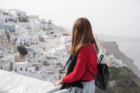 Young asian girl in red sweater and black backpack enjoy the landscape view while on vacation in Santorini, Greece.の写真素材