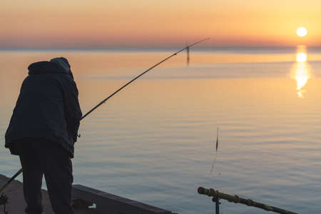 Silhouette of adult man fishing at the Pier Geelong against calming ocean with sunrise during golden hourの写真素材