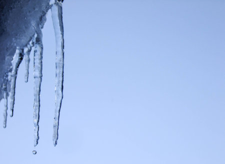Two Icicles and falling Water Drop from one of them on the Roof in blue Sky Background