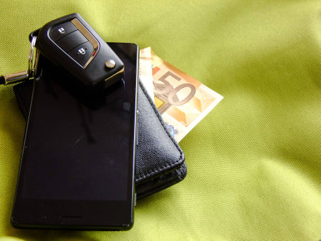 Black Car Keys with small Pendant on top of Smartphone on top of Black Wallet with fifty Euro in greenish Textile Backgroundの写真素材