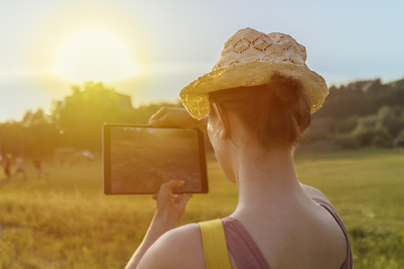 Young girl in a hat takes photo or video of nature with tablet computer. Back view. Warm sunset colours, soft sunlight. Tourist woman on vacation.の写真素材