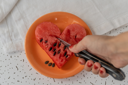 Woman hand holds a knife and cutting the Heart made of watermelon on small orange plate, white background with small black texture, selective focusの写真素材