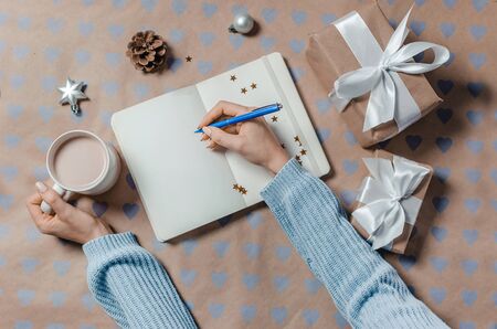 Female writing wish list in to notebook near christmas gifts