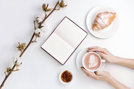 Hands holding coffee with latte art and notebook with croissant, jam and cotton branchの写真素材