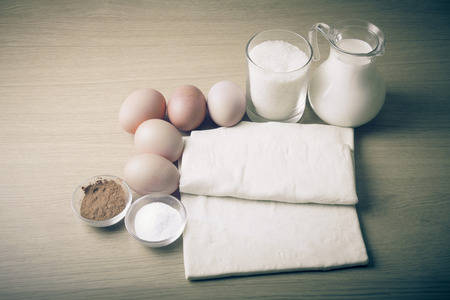 Milk, sugar, cocoa, puff dough, eggs and salt on a wooden board. Ingredients for pastry. Toned.の写真素材