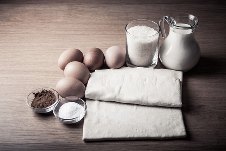 Milk, sugar, cocoa, puff dough, eggs and salt on a wooden board. Ingredients for pastry. Toned.の写真素材