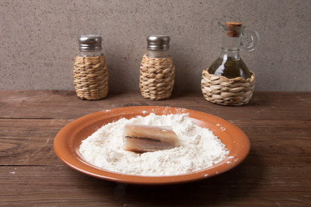 Pepper, salt, olive oil and one piece of fresh fish with flour on a plate on a wooden table.の写真素材
