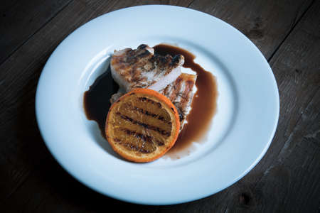Grilled meat with orange on white ceramic plate on old wooden table. Toned.の写真素材
