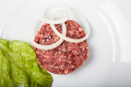 Raw cutlet of minced meat with leaf of salad and rings of onion on a white plate.の写真素材