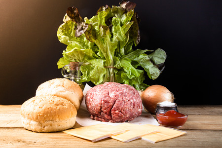Meat, cheese, onions, herbs, salt, ketchup on a wooden table over black background. Ingredients for a hamburger. Toned.の写真素材