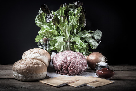 Meat, cheese, onions, herbs, salt, ketchup on a wooden table over black background. Ingredients for a hamburger. Toned.の写真素材