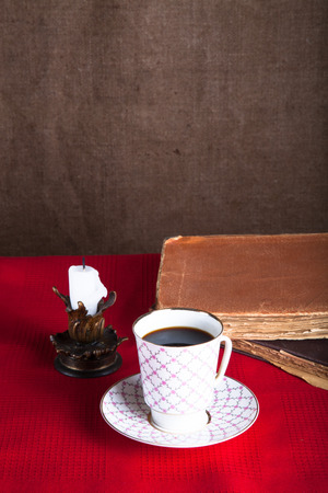 Stack of old books, cup of coffee, and ancient candle on the red tablecloth. Burlap background.の写真素材