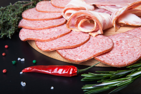 Slices of sausage and bacon on a round cutting board with rosemary, thyme, colored and chili pepper...