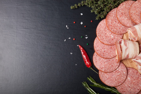 Slices of sausage and bacon on a round cutting board with rosemary, thyme, colored and chili pepper and sea salt on a black background.の写真素材
