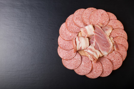 Slices of sausage and bacon on a round cutting board on a black background.の写真素材