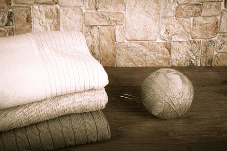 Stack of knitting clothes and ball of yarn with knitting needles on wooden table opposite a stony wall. Toned.の写真素材