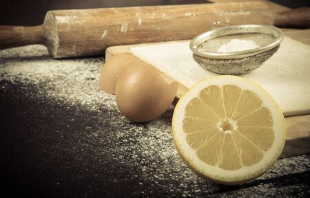 Set for home baking on a black background with flour. Rolling pin, dough, half of lemon, baking form, eggshell. Toned.の写真素材