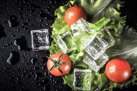 Red cherry tomatos, green salad and ice cubes on black wet table. Selective focus. Toned.の写真素材