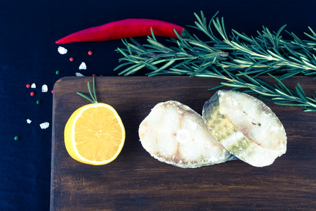 Fish steaks, rosemary, red chili pepper, half a lemon and sea salt on brown cutting board on a black...