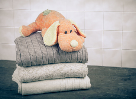 Knitted toy dog on the stack of knitted clothes on wooden table opposite a light tile wallの写真素材