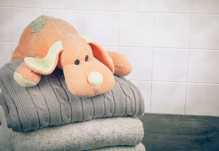 Knitted toy dog on the stack of knitted clothes on wooden table opposite a light tile wall. Toned.の写真素材