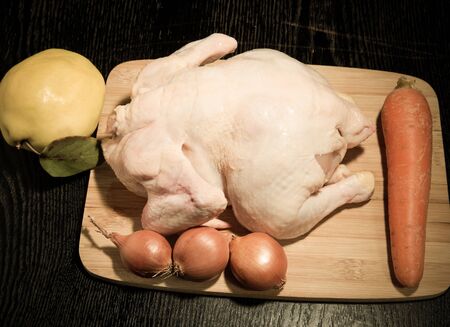 Fresh whole chicken with fruit and vegetables is prepared for cooking on a light wooden cutting board on a black table. Toned.の写真素材