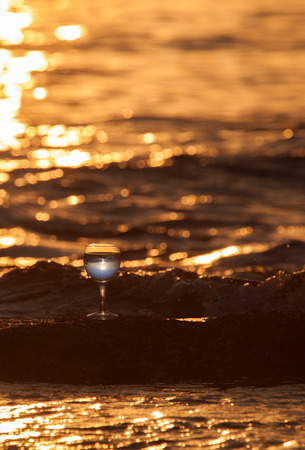 Glass of white vine with reflections of sun and sea on the background of beautiful sunset. Selective focus.の写真素材