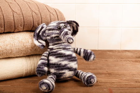 Knitted toy elephant with the stack of knitted clothes on wooden table opposite a light tile wall. Toned.の写真素材