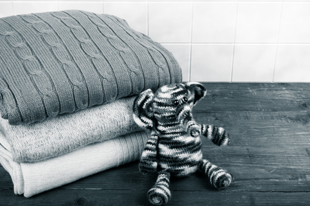 Knitted toy elephant with the stack of knitted clothes on wooden table opposite a light tile wall. Toned.の写真素材