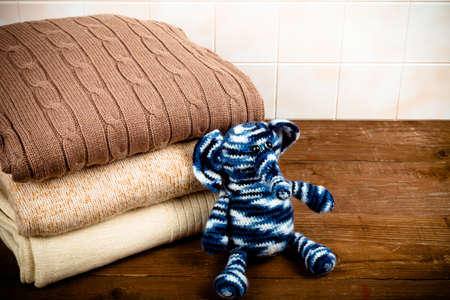 Knitted toy elephant with the stack of knitted clothes on wooden table opposite a light tile wall. Toned.の写真素材