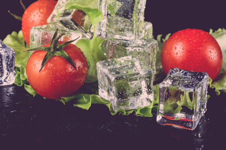 Banch of red cherry tomatos, green salad and ice cubes on black wet table. Selective focus. Toned.の写真素材