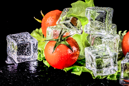 Banch of red cherry tomatos, green salad and ice cubes on black wet table. Selective focus. Toned.の写真素材