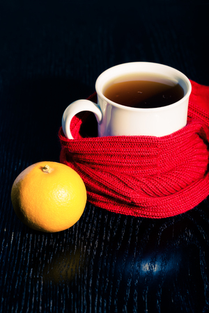 White mug with tea and warm knitted scarf on the dark background. Toned.