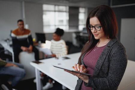An attractive and smiling young brazilian businesswoman using digital tablet while her colleague sitting in background at officeの写真素材