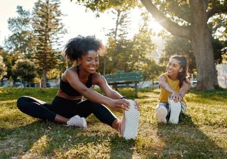 Young diverse female friends sitting on green grass stretching her legs in the morning sunlight at park - diverse friends warming up before doing group exerciseの写真素材