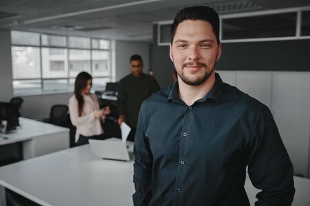 Portrait of a professional young businessman smiling and looking at camera while other business people talking in background at officeの写真素材