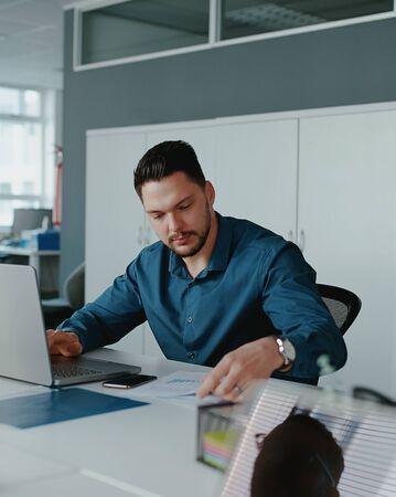 A young male entrepreneur using laptop computer working on reports sitting at office deskの写真素材