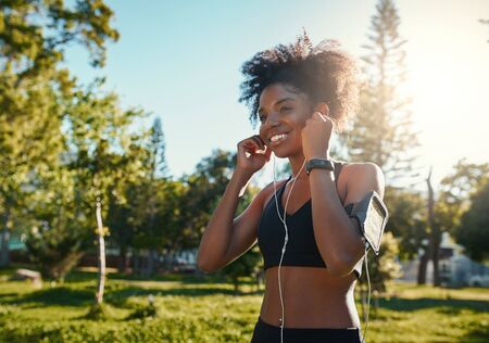 Happy young black african american woman enjoying listening to music on earphones while jogging workout in the bright sunlight at parkの写真素材