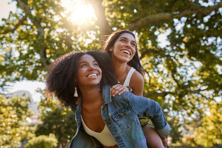 Two diverse multiracial friends having fun laughing and giving a piggyback to each other in the park on a warm summer dayの写真素材
