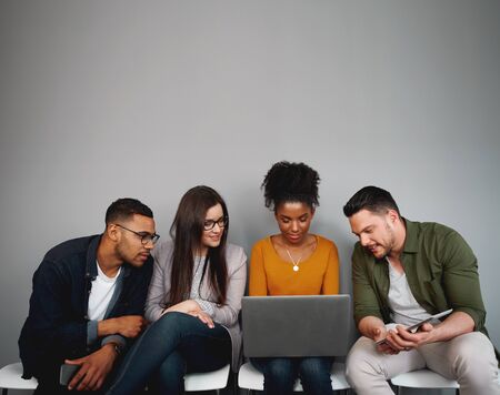 Group of diverse friends sitting on chairs in queue watching videos online or using social media app...